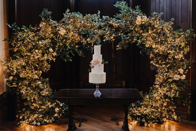 Luxury 4 tier white wedding cake with vertical ruffles and sugar flowers surrounded by a floral, fairy-lit arch | The Elvetham Hampshire | Melissa Megan Photography | Louise Hayes Cake Design