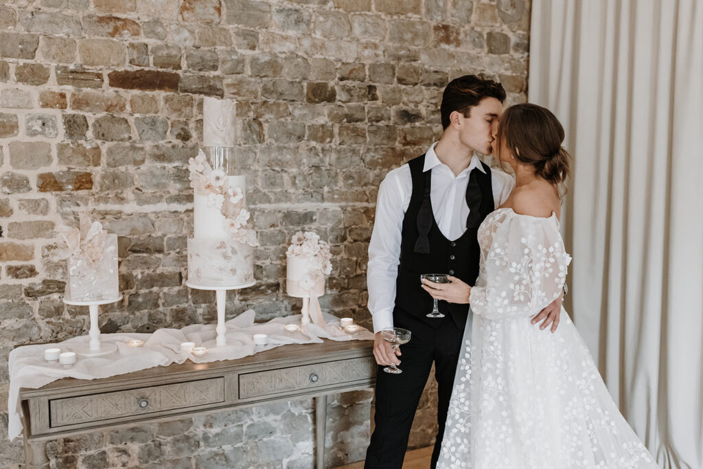 Bride and groom kissing with champagne and a trio of nude, blush contemporary wedding cakes with wafer paper sails and sugar flowers | Louise Hayes Cake Design | Melissa Megan Photography | Cowdray Wedding